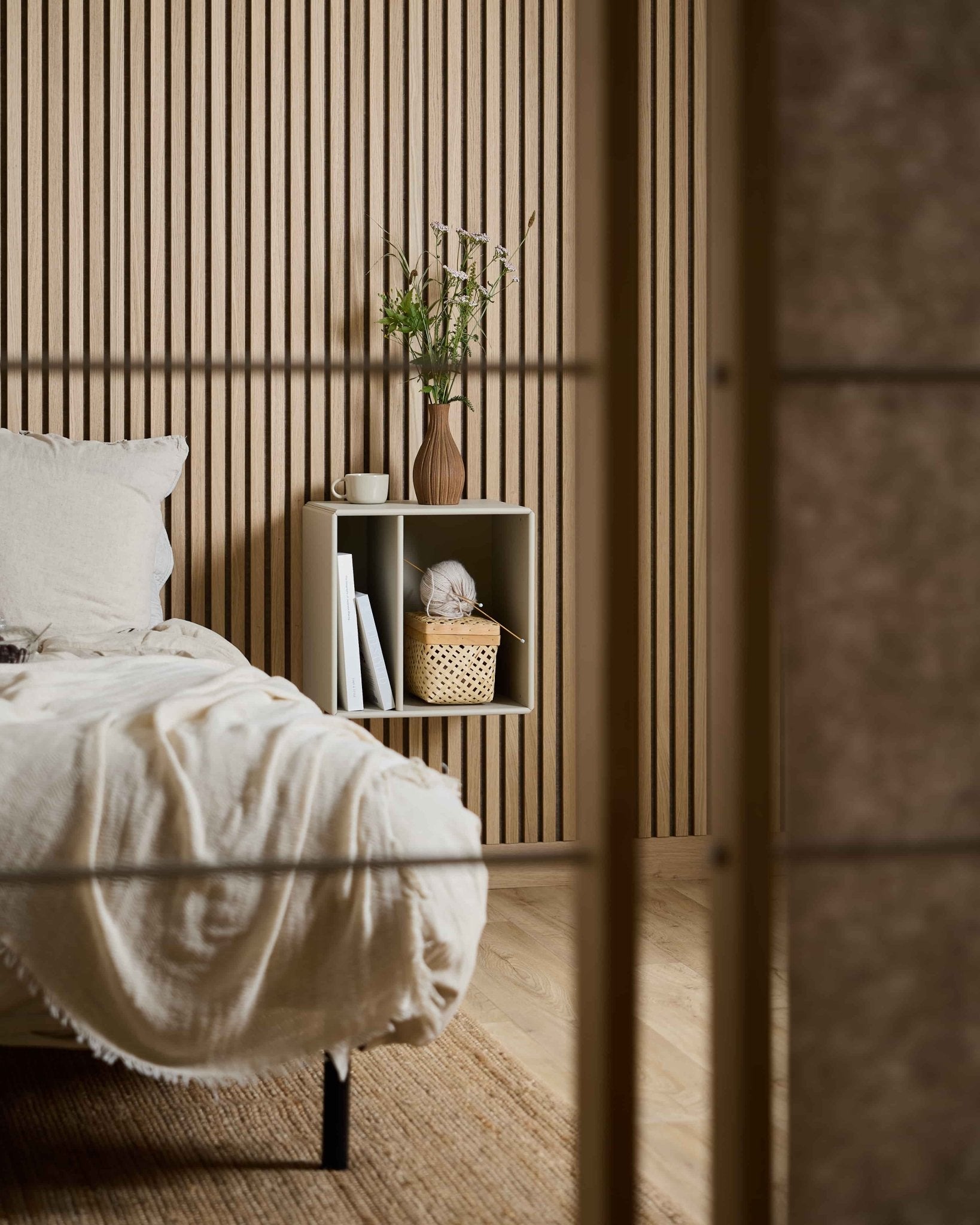Camera da letto in stile minimalista con libreria WoodUpp Arka color beige, libri, cesto e vaso di fiori, il tutto sistemato davanti a un pannello acustico a doghe Akupanel in stile nordico.